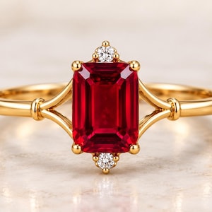 May include: A gold ring featuring a central emerald-cut red gemstone held by four prongs. Two small, round, clear stones are set vertically above and below the main red stone. The band splits into a dual-arm design where it meets the setting.