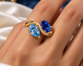 May include: A gold bypass ring featuring two pear-cut gemstones, one light blue and one dark blue, set in a polished gold band with a curved design accented by a row of small white stones along the center bridge of the ring setting.