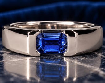 May include: A polished silver-toned metal ring featuring a single emerald-cut blue sapphire gemstone set horizontally in a tension-style setting. The band has a smooth, wide, and solid construction with a reflective finish.