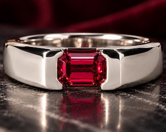 May include: A polished silver-toned metal ring featuring a single rectangular emerald-cut red gemstone set horizontally in the center of a wide, smooth band with a clean, minimalist design and a reflective metallic finish.