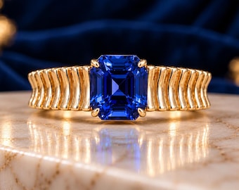 May include: A gold ring featuring a central emerald-cut blue gemstone held by four prongs. The band is constructed with a series of vertical, curved, ribbed gold segments that extend from the center stone around the finger.
