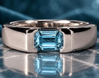 May include: A polished silver-toned metal ring featuring a rectangular, emerald-cut light blue gemstone set securely in the center. The band has a smooth, wide, and solid construction that frames the faceted blue stone horizontally.