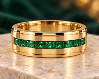 May include: A polished gold band ring featuring a central channel set with a continuous row of square-cut vibrant green gemstones. The metal surface is smooth and reflective, showcasing a wide profile with raised edges framing the stones.