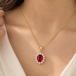 May include: A gold necklace featuring an oval-cut red gemstone pendant surrounded by a halo of small, round, clear white stones. The pendant is suspended from a delicate gold chain, creating a classic and elegant piece of jewelry.