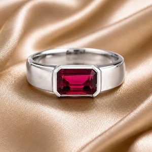 Könnte beinhalten: Ein silberfarbener Ring mit einem rechteckigen, smaragdgeschliffenen, tiefroten Edelstein, der in eine glatte, polierte Metallschiene eingefasst ist. Der Stein ist in einer Zargenfassung sicher gehalten, was dem Schmuckstück ein klares Design verleiht.