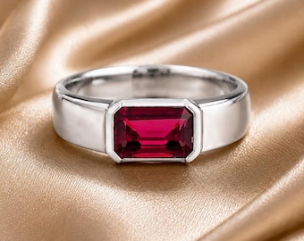 Peut inclure: Une bague argentée ornée d'une pierre précieuse rectangulaire rouge foncé taille émeraude, montée sur un anneau en métal lisse et poli. La pierre est maintenue par un serti clos, offrant un aspect net et uniforme à ce bijou.