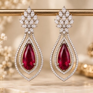 May include: A pair of drop earrings featuring a large pear-shaped deep red gemstone at the centre, surrounded by two concentric teardrop-shaped frames encrusted with small clear stones, topped with a cluster of marquise-cut clear stones.