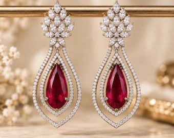May include: A pair of drop earrings featuring a large pear-shaped deep red gemstone at the center, surrounded by two concentric teardrop-shaped frames encrusted with small clear stones, topped with a cluster of marquise-cut clear stones.