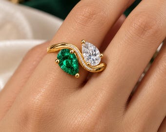 May include: A gold bypass ring featuring two pear-cut stones, one vibrant green and one clear white, set in a polished gold band with a delicate row of small white accent stones along the curved bridge connecting the two main gems.