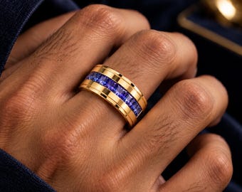 May include: A wide gold band ring featuring a central channel set with a row of square-cut, vibrant purple-blue gemstones. The polished gold metal has a smooth, reflective surface that frames the continuous line of faceted stones.