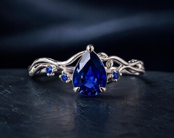 May include: A silver-toned ring featuring a large pear-shaped blue center stone held by prongs. The band is designed with a vine-like motif, incorporating small leaves and four smaller round blue accent stones flanking the central pear-shaped gem.