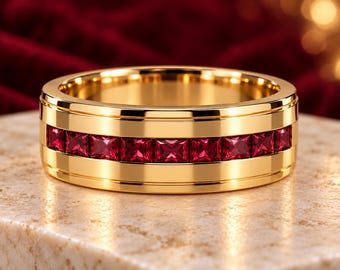 May include: A polished gold band ring featuring a channel setting with a continuous row of square-cut, deep red rubies encircling the center. The ring has a smooth, reflective metallic finish and a wide, flat profile with raised edges.