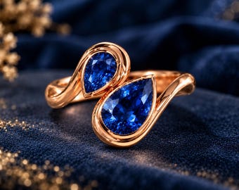 May include: A bypass ring crafted from rose gold, featuring two pear-cut deep blue sapphires set in smooth, polished metal bezels. The design showcases a continuous, flowing band that curves around the finger, highlighting the vibrant blue stones.