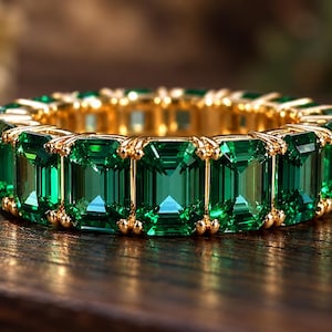 Peut inclure: Une bague alliance éternité composée d'une rangée continue de pierres vertes taille émeraude serties dans une monture en métal doré poli. La bague présente une série de gemmes rectangulaires à facettes disposées côte à côte en cercle.