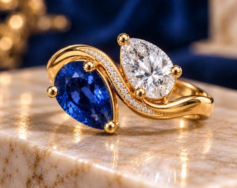 May include: A gold bypass ring featuring a pear-cut blue sapphire and a pear-cut clear white gemstone. The band is accented with a row of small pavé-set white stones along the curved bridge connecting the two primary pear-shaped gems.