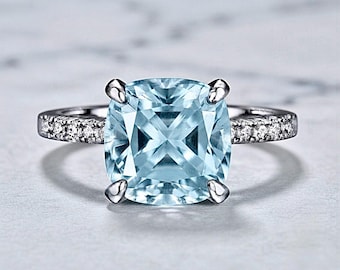 May include: A silver-toned ring featuring a large, cushion-cut, light blue gemstone held by four prongs. The band is accented with a row of small, clear, round-cut stones embedded along each side of the central setting.