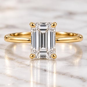Peut inclure: Une bague en or avec une grande pierre précieuse transparente taille émeraude montée sur quatre griffes. L'anneau est en or jaune poli et lisse, et la pierre centrale présente une forme rectangulaire avec des facettes qui reflètent la lumière.