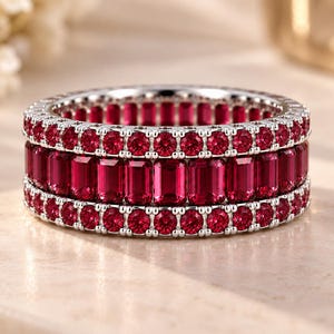 May include: A silver-toned band ring featuring a central row of emerald-cut red stones flanked by two outer rows of smaller, round-cut red stones. The metal setting is polished and holds the stones in a continuous, full-eternity style arrangement.