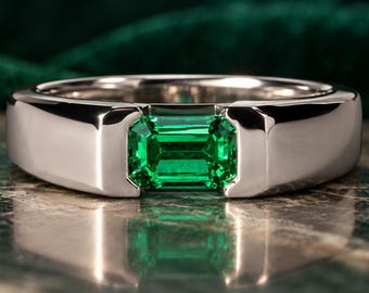 May include: A polished silver-toned band ring featuring a single emerald-cut green gemstone set horizontally in a tension-style setting. The metal band has a smooth, reflective surface and a wide, solid construction that frames the vibrant green stone.