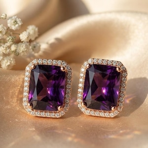May include: A pair of rectangular stud earrings featuring large, deep purple faceted stones set in rose gold metal. Each central stone is surrounded by a halo of small, clear, sparkling white stones, creating a bright and elegant contrast.