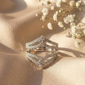 May include: A silver-toned ring guard featuring two chevron-shaped bands connected at the base. The bands are encrusted with numerous small, clear, sparkling stones along their entire V-shaped surfaces, creating a layered, symmetrical appearance.