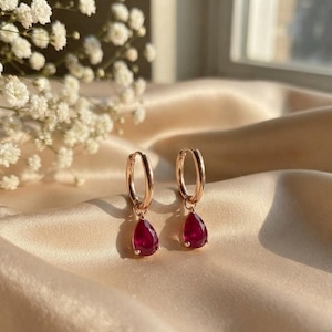 May include: A pair of rose gold hoop earrings featuring dangling teardrop-shaped deep red gemstones. The earrings have a polished metallic finish and a secure hinged closure, showcasing a vibrant crimson hue in the faceted stone drops.