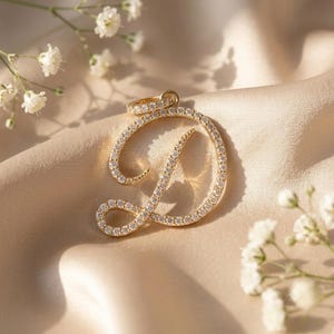 Pode incluir: Um pingente dourado em forma de letra D cursiva, incrustado com pequenas pedras transparentes em toda a sua superfície. O pingente apresenta um pequeno aro circular no topo para fixação e possui um acabamento metálico polido e reflexivo.