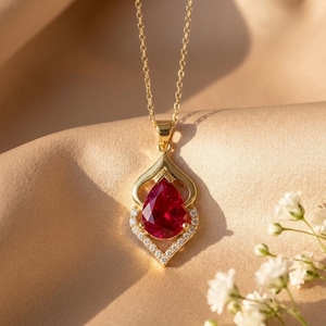May include: A gold necklace featuring a pear-shaped red gemstone pendant set in a decorative gold frame accented with small clear stones. The pendant hangs from a delicate gold chain, creating a refined and elegant piece of jewelry.