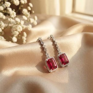May include: A pair of drop earrings featuring rectangular, deep red gemstones set in silver-toned metal. Each earring has a vertical row of small, clear, round stones leading to the larger red stone, which is surrounded by a halo of clear stones.