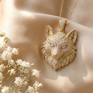 Könnte beinhalten: Ein goldfarbener Anhänger in Form eines Fuchskopfes, der vollständig mit kleinen klaren Steinen besetzt ist und an einer passenden goldfarbenen Kette hängt. Der Anhänger zeigt detaillierte Gesichtszüge und spitze Ohren mit einer Öse.