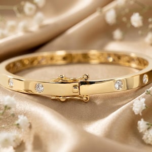 Op de afbeelding: Een gepolijste gouden armband met een reeks ronde, heldere, gefacetteerde stenen die met tussenpozen langs de band zijn geplaatst. De armband is voorzien van een veilige scharnierende doosluiting met een veiligheidsklepje voor een sluitend geheel.