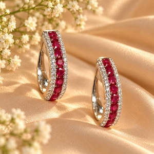 May include: A pair of hoop earrings featuring a central row of round, vibrant red rubies flanked by two rows of small, clear white diamonds set in polished silver-toned metal, creating a curved, open-frame design with a secure hinged closure.
