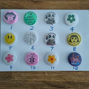 May include: A collection of twelve circular pin-back badges arranged in three rows of four. Designs include a mouth with a tongue, a green circle with text, a panda on a skateboard, a green flower, a yellow smiley face, a tree, a sun face, and mushrooms.