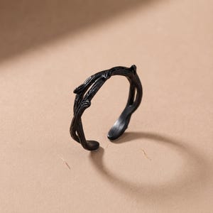 May include: A matte black ring featuring a textured design that mimics the appearance of intertwined tree branches. The band is open at the front, showcasing a detailed, organic surface with small knots and ridges throughout the metal structure.