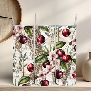 May include: A cylindrical tumbler with a straw featuring a pattern of red cherries, green leaves, white cherry blossoms, and tan leopard print spots on a cream background. The design wraps around the entire surface of the tall beverage container.