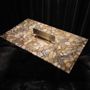 May include: A rectangular coffee table featuring a tabletop composed of numerous polished, multi-coloured agate slices in shades of beige, tan, grey, and white. A small gold-toned metal sign with the text AlphaLuxDesigns sits on the surface.