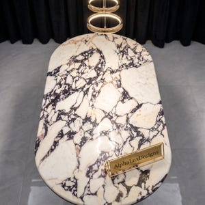 May include: An oval dining table with a white marble top featuring prominent dark purple and black veining. A gold-toned metal nameplate with the text AlphaLuxDesigns sits on the surface. Above the table hangs a three-tiered circular gold light fixture.