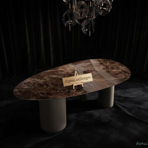 Puede incluir: Una mesa de comedor ovalada con una superficie de mármol marrón pulido con vetas naturales y dos bases cilíndricas sólidas de color gris claro. Un pequeño caballete de madera sobre la mesa muestra un letrero que dice AlphaLuxDesgins.
