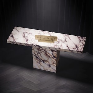 Puede incluir: Una mesa consola rectangular con un patrón de mármol blanco y morado y un acabado liso. La mesa tiene una superficie ancha sostenida por una base de pedestal central única. Una pequeña placa rectangular dorada muestra el texto AlphaLuxDesigns.