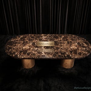 Puede incluir: Una mesa de comedor ovalada con una superficie de mármol marrón oscuro con vetas blancas intrincadas y dos bases cilíndricas de madera clara. Una pequeña placa rectangular dorada con el texto AlphaLuxDesigns está centrada en la mesa.