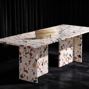 Op de afbeelding: Een rechthoekige eettafel met een wit terrazzo oppervlak met veelkleurige steensplinters in tinten bruin, blauw en beige. De tafel wordt ondersteund door twee brede, bijpassende terrazzo plaatpoten en is voorzien van een gouden plaatje met AlphaLuxDesigns.