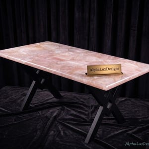 May include: A rectangular table featuring a polished, translucent pale pink rose quartz slab top with a natural crystalline texture. The table is supported by a black metal base with an X-shaped frame design. A gold nameplate reads AlphaLuxDesigns.