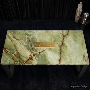 Op de afbeelding: Een rechthoekige tafel met een gepolijst groen onyx stenen blad met opvallende witte en bruine aders. De tafel wordt ondersteund door donkere metalen poten en heeft in het midden een klein goudkleurig plaatje met de tekst AlphaLuxDesigns.