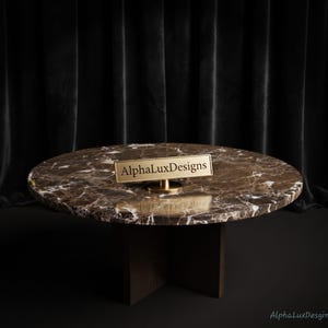 Pode incluir: Uma mesa de centro redonda com tampo de mármore castanho com veios brancos e base de madeira escura. Uma pequena placa retangular de metal dourado com suporte está colocada na superfície, exibindo o texto preto AlphaLuxDesigns.