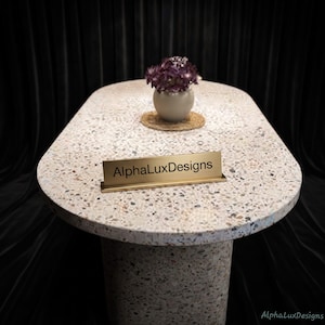 May include: An oval-shaped table with a speckled terrazzo surface and a matching pedestal base. A small, round, light-coloured vase containing purple flowers sits on a circular woven mat. A gold-toned metal nameplate displays the text AlphaLuxDesigns.