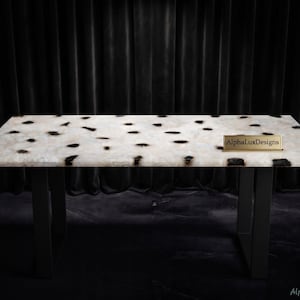 May include: A rectangular table featuring a white, translucent stone surface with scattered dark brown and black organic spots. The table is supported by two black metal U-shaped legs. A small gold-toned plaque on the surface reads AlphaLuxDesigns.