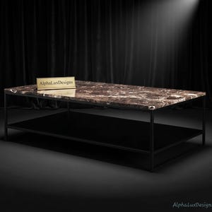 Op de afbeelding: Een rechthoekige salontafel met een gepolijst bruin marmeren blad met witte aders en een zwart metalen frame met een onderste plank. Een klein goudkleurig rechthoekig plaatje met de tekst AlphaLuxDesigns ligt op het marmeren oppervlak.