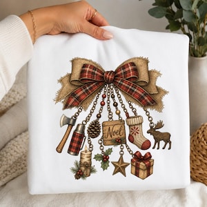 May include: A white sweatshirt featuring a graphic of a large plaid bow with hanging chains attached to various charms including an axe, a thermos, a pinecone, a wooden sign reading Noel, a stocking, a moose, a candle, holly, a star, and a gift.