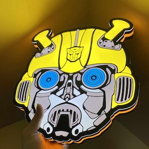 Puede incluir: Un letrero de pared iluminado en amarillo y gris con la forma de la cabeza del personaje Bumblebee de Transformers. El letrero presenta ojos circulares de color azul brillante, un logotipo de Autobot central en la frente y una luz de borde naranja.