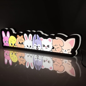 Puede incluir: Un letrero luminoso LED rectangular que presenta una fila de ocho personajes animales de dibujos animados coloridos con expresiones distintas. El letrero tiene un borde negro y emite un brillo suave, mostrando un conejo rosa y un pollito amarillo.